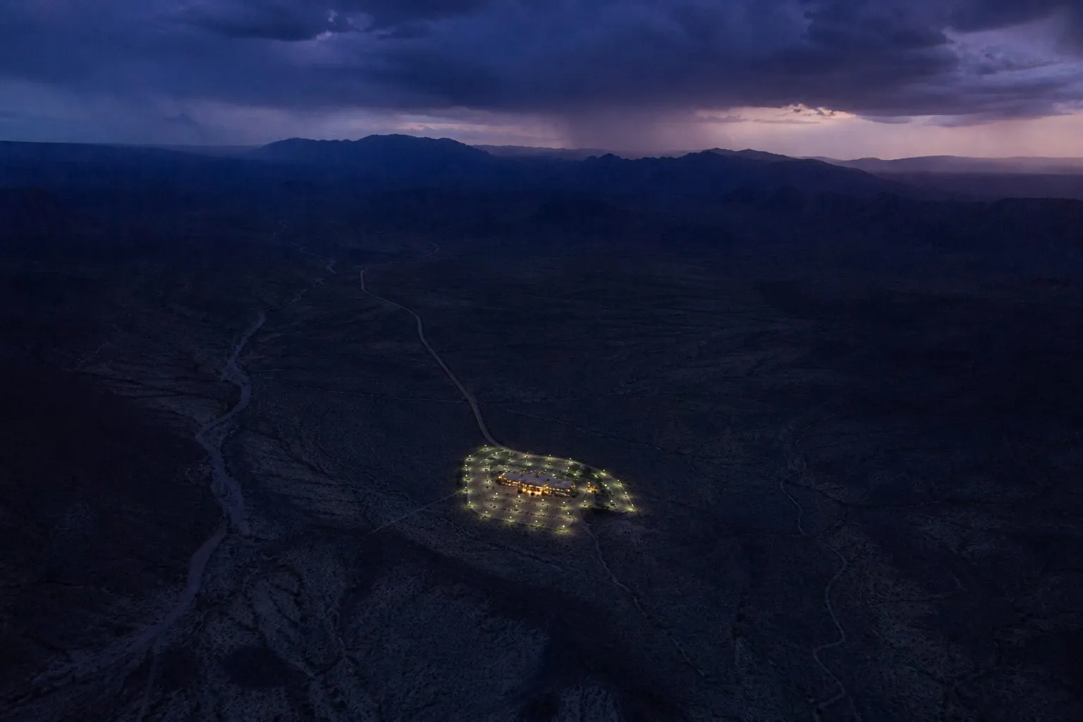 Aerial night view of a single lit complex isolated in dark desert terrain, one road arriving from the distance